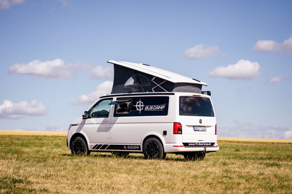 Weißer VW T6.1 mit geöffnetem Aufstelldach auf einer Wiese unter blauem Himmel